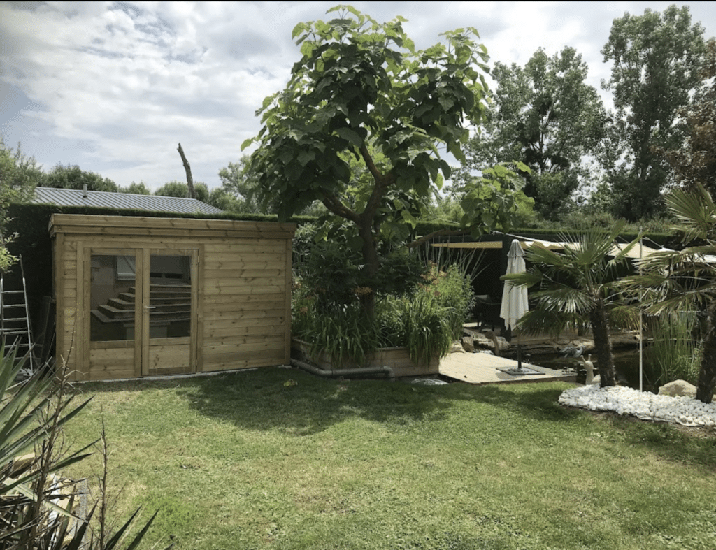 Abri de jardin bois avec baie vitrée installé au bord d'un bassin paysager arboré