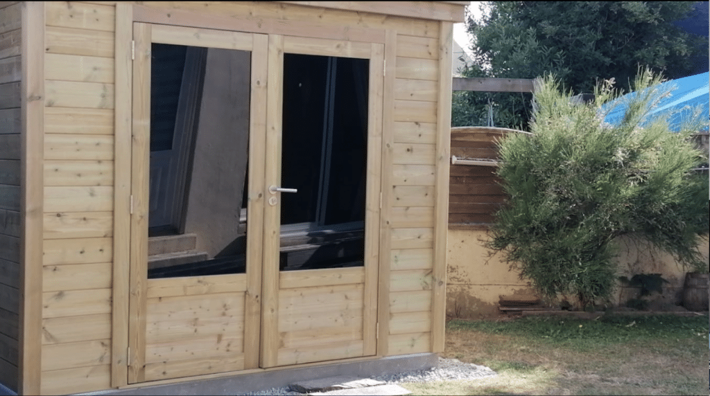 Abri jardin en bois avec double porte vitrée, installation client satisfaite dans un environnement verdoyant