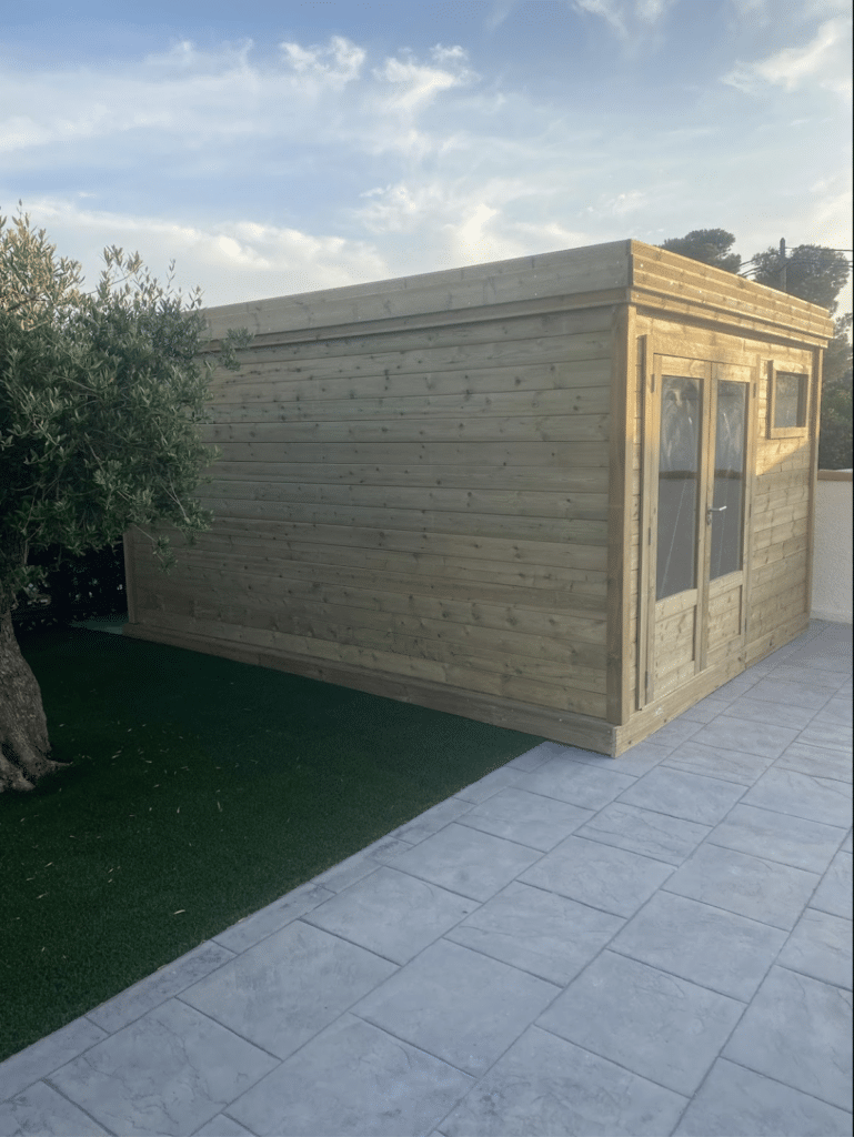 Abri de jardin en bois à toit plat avec porte vitrée et fenêtre, posé sur terrasse carrelée près d'un olivier