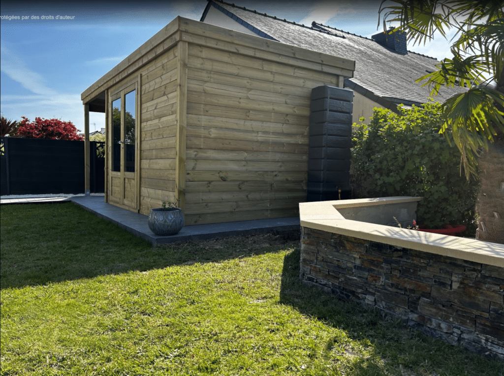 Abri de jardin en bois massif installé dans un jardin verdoyant, avec terrasse et mur de soutènement en pierre